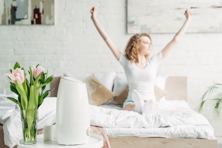 Im Hintergrund sitzt eine Frau auf einem Bett und reckt die Arme in die Höhe. Im Vordergrund ist ein Luftreiniger zu sehen.