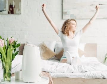 Im Hintergrund sitzt eine Frau auf einem Bett und reckt die Arme in die Höhe. Im Vordergrund ist ein Luftreiniger zu sehen.