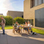 Eine Familie grillt im Garten, im Hindergrund an der Hausfassade ist eine Minisolaranlage zu sehen