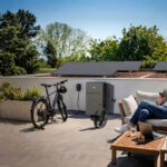 Mann mit Hund sitzt vor einer Mini-Solaranlage, an deren Speicher ein E-Bike und Ventilator angeschlossen sind