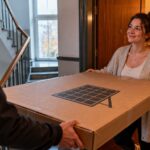 Paketbote übergibt einer Frau an der Wohnungstür ein großes Paket mit einem Solarmodul