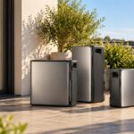 Drei silbergraue Batteriespeicher unterschiedlicher Größe stehen bei Sonnenschein auf einer gefliesten Dachterrasse neben Kübelpflanzen, im Hintergrund die Dächer einer Stadt