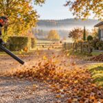 Mann mit Gehörschutz bläst mit einem Benzin-Laubbläser Herbstlaub von einem Kiesweg auf einem großen Grundstück