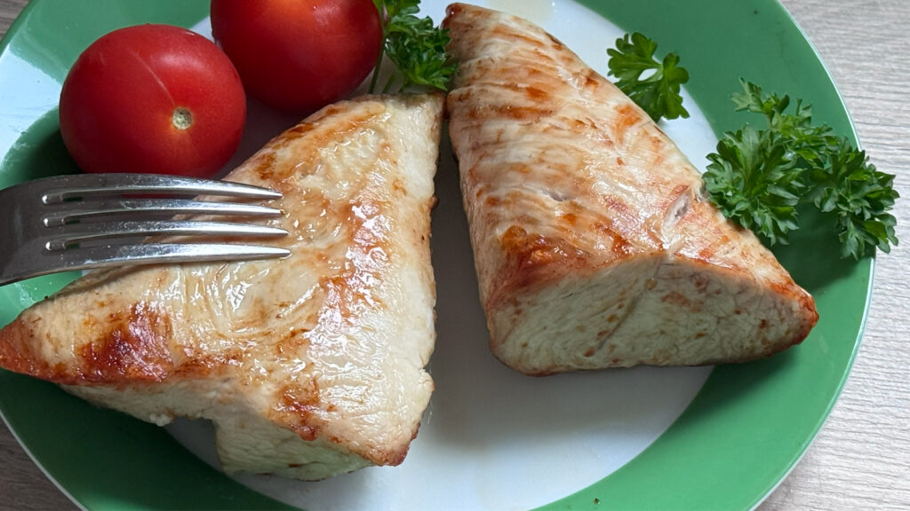 putenbrust-im-grillmodus-gegart Gabel berührt ein gebräuntes Stück Putenbrust, daneben liegen Petersilie und Tomaten als Garnitur