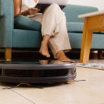 Eine Frau sitzt mit ihrem Laptop auf dem Sofa, davor reinigt ein Roboter den Boden